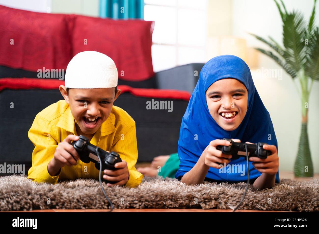 Two muslim sibling kids playing videogame using joystick while lying on floor at home - Concept of children unhealthy playing position and gaming Stock Photo