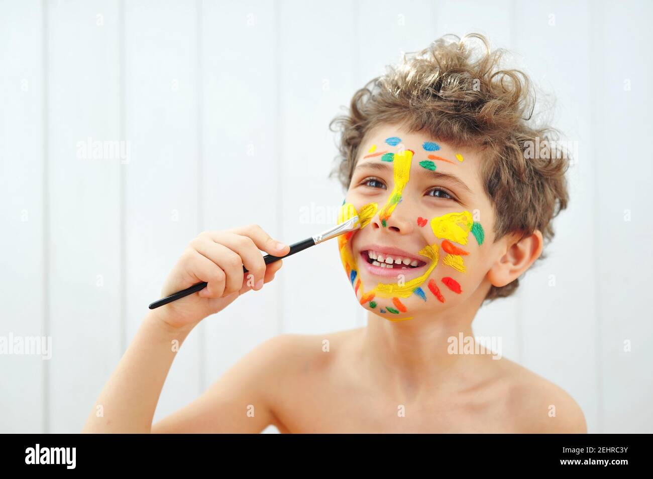 Happy little boy holds a paintbrush in his hands and draws colorful stripes on his face. Stock Photo