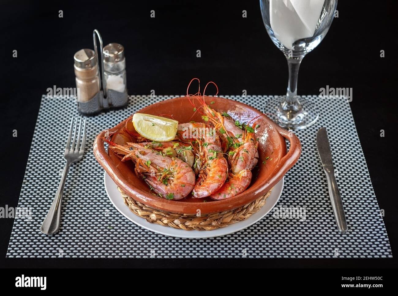 Cooked shrimp inside a clay bowl, cutlery on the sides and a glass of water. Composition of a meal ready to be eaten. Stock Photo