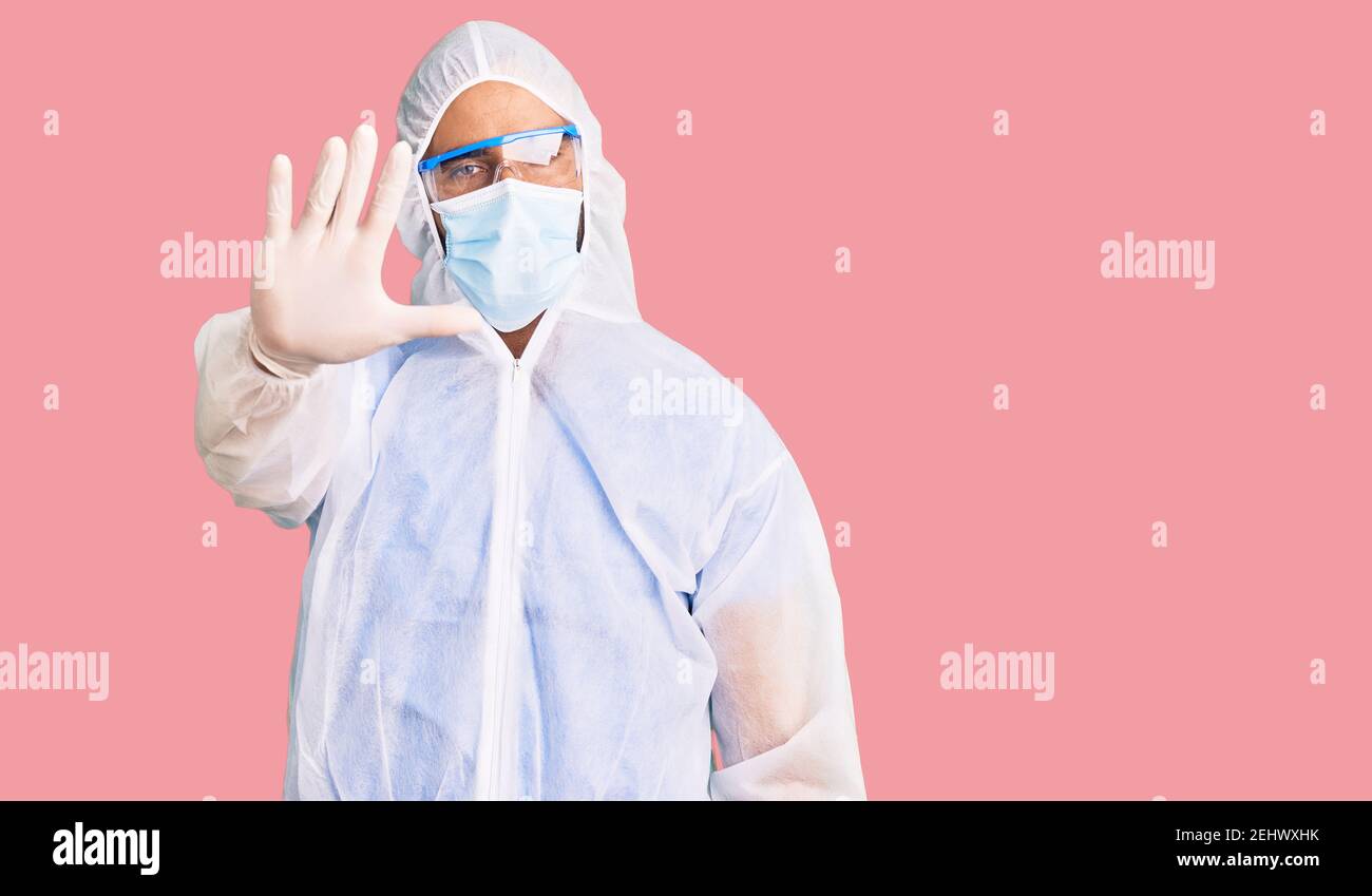 Young hispanic man wearing doctor protection coronavirus uniform and medical mask doing stop sing with palm of the hand. warning expression with negat Stock Photo