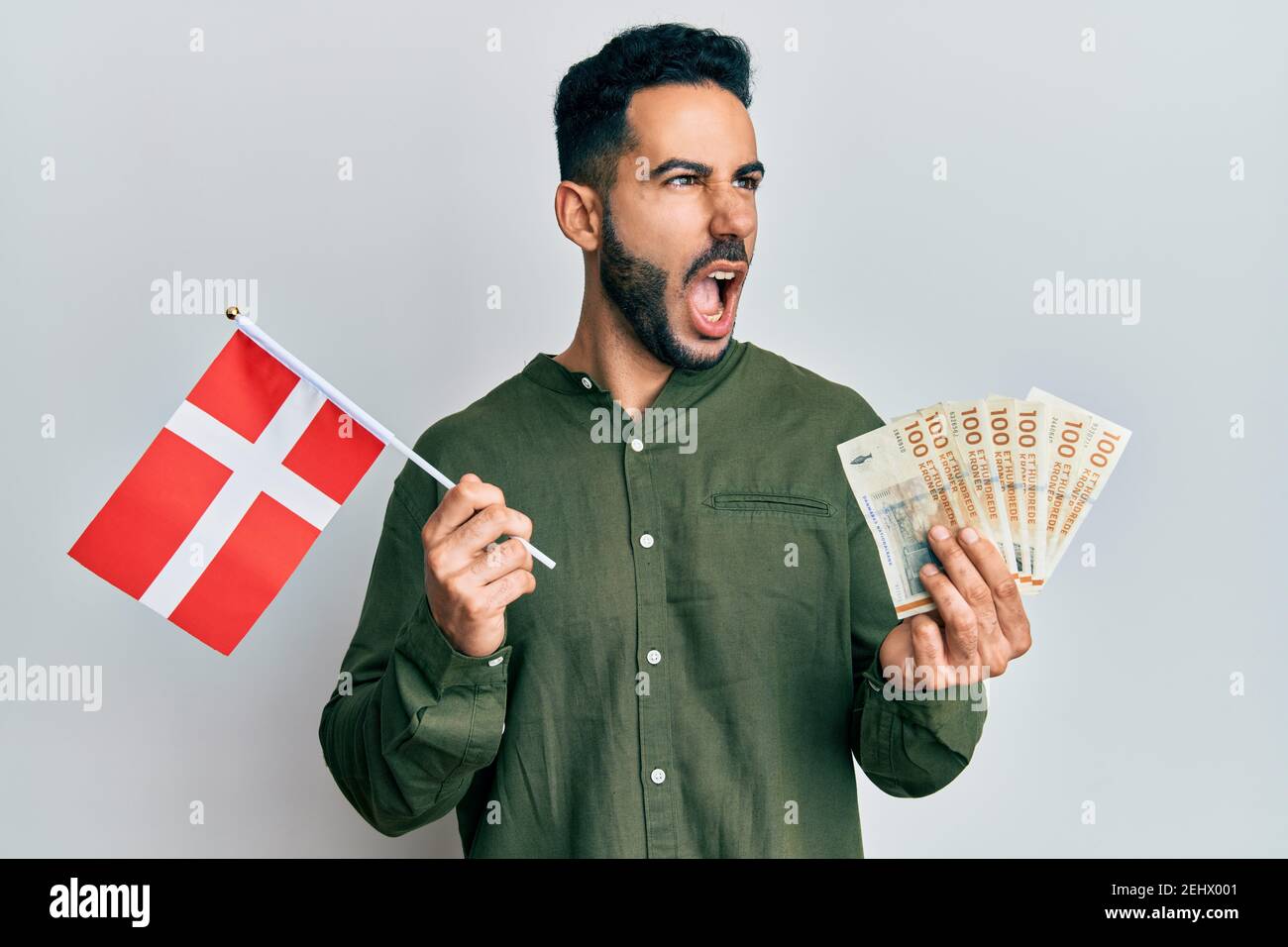 Young hispanic man holding denmark flag and krone banknotes angry and ...