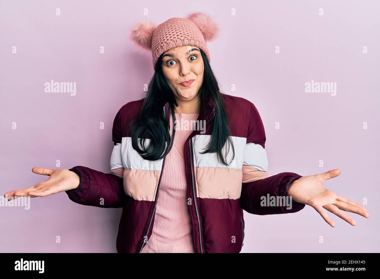 Young hispanic woman wearing wool winter sweater and cap clueless and confused expression with arms and hands raised. doubt concept. Stock Photo