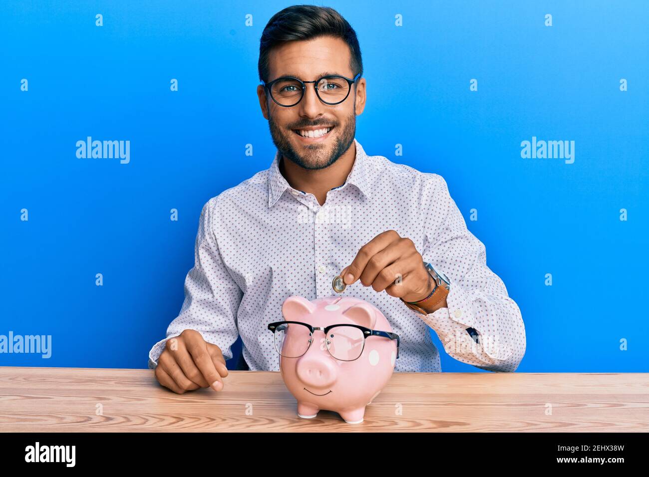 Handsome hispanic man holding piggy bank with glasses looking positive ...