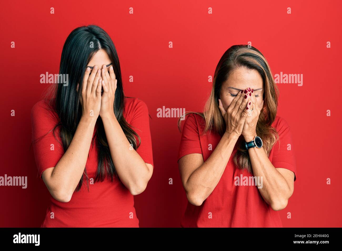 Hispanic family of mother and daughter wearing casual clothes over red background with sad expression covering face with hands while crying. depressio Stock Photo