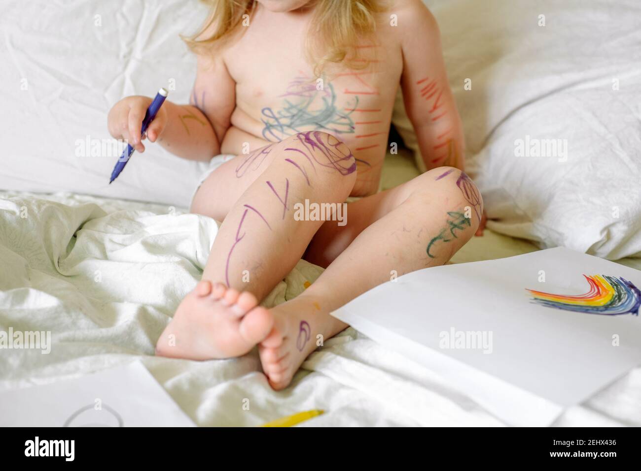 girl draws a rainbow on paper with felt-tip pens on bed. children play in the morning at home. mischievous naughty baby, smeared hands, feet and face Stock Photo