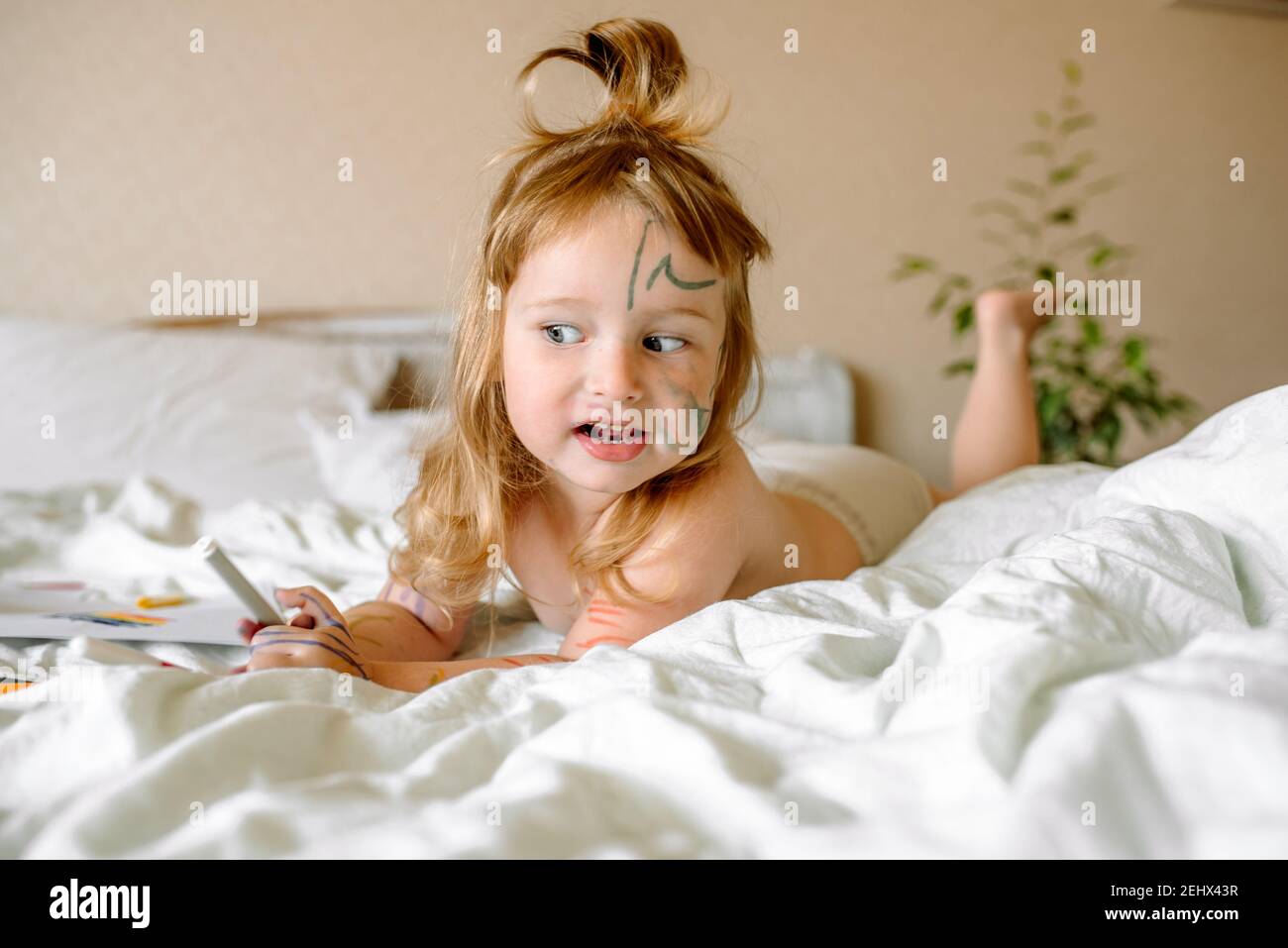girl draws a rainbow on white paper with felt-tip pens on the bed. children play in the morning at home. mischievous naughty baby, smeared hands, feet Stock Photo
