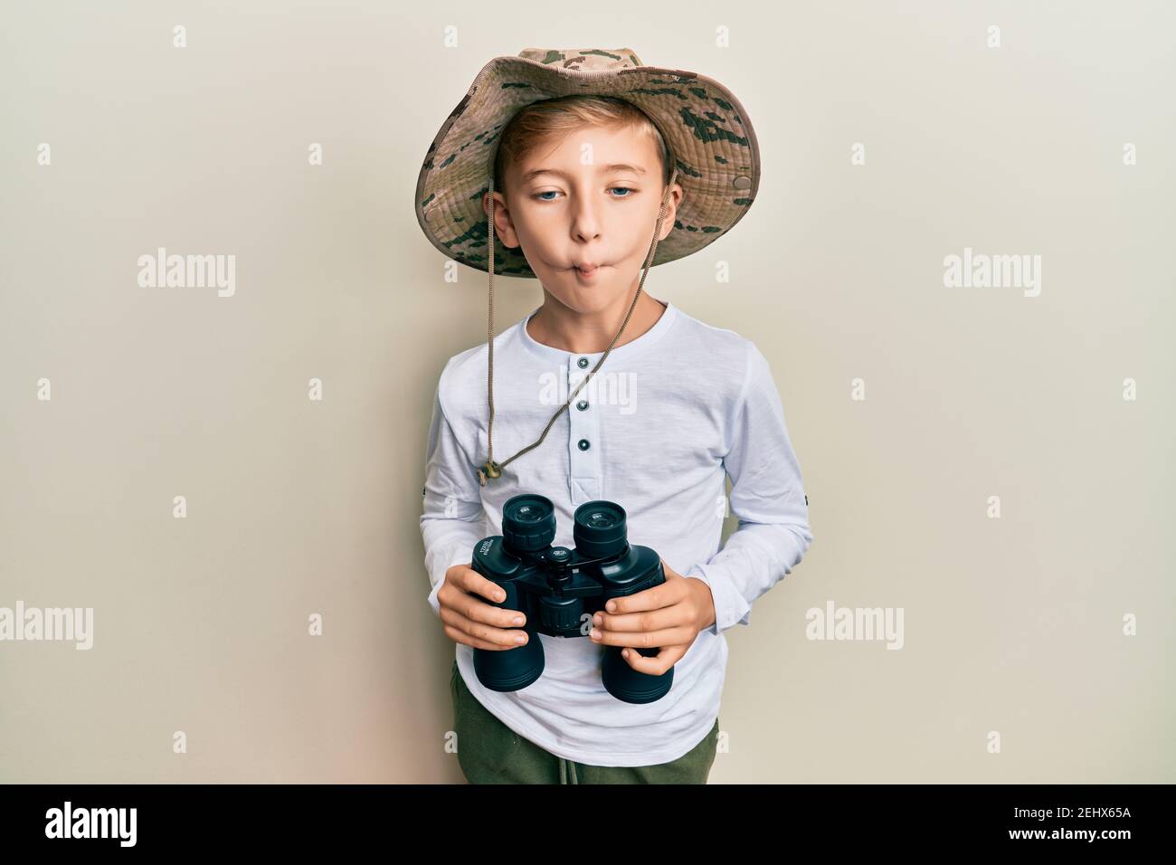 Little caucasian boy kid wearing explorer hat holding binoculars making fish  face with mouth and squinting eyes, crazy and comical Stock Photo - Alamy