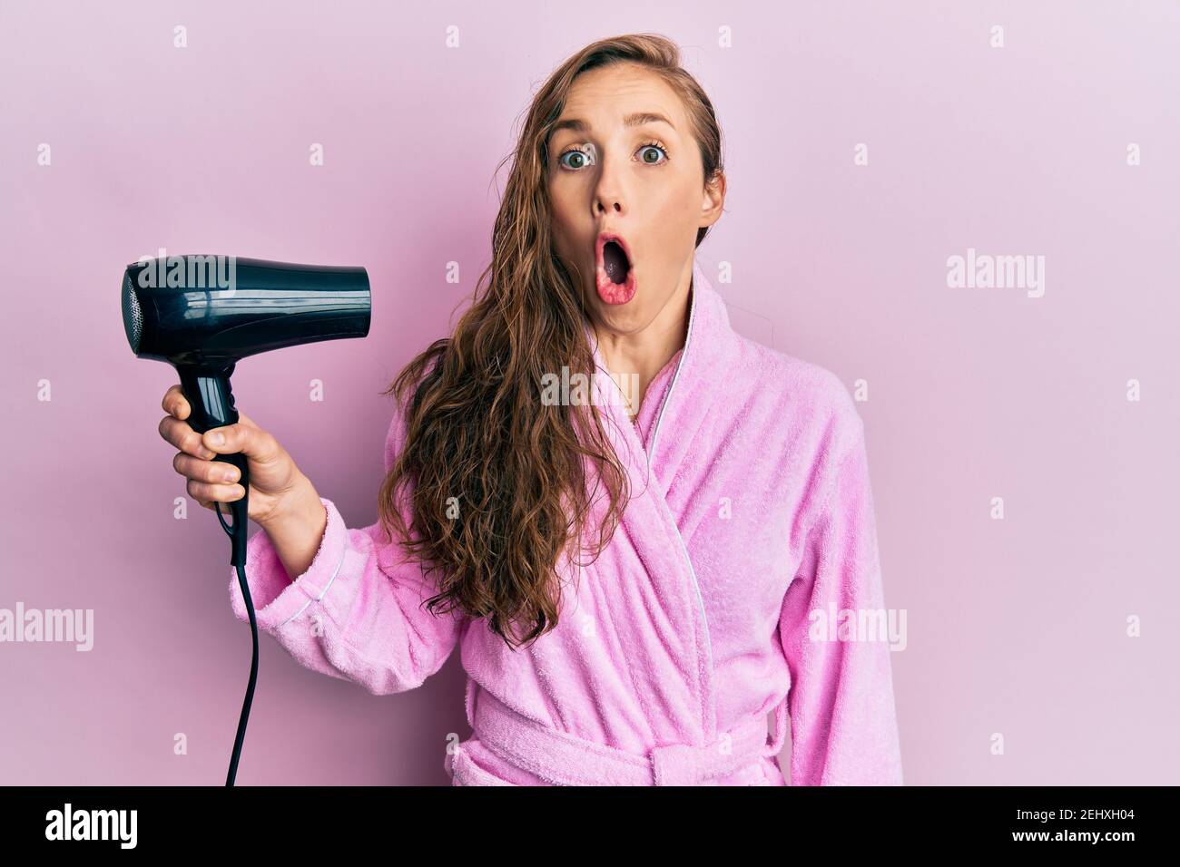 Young blonde woman wearing bathrobe using hair dryer scared and amazed