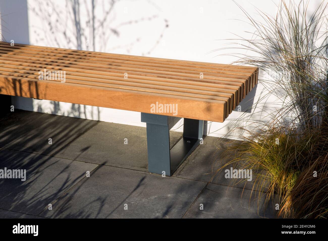 A modern wooden garden wood bench with metal frame legs seating area on a dark stone paved patio with white wall and ornamental grasses England UK GB Stock Photo