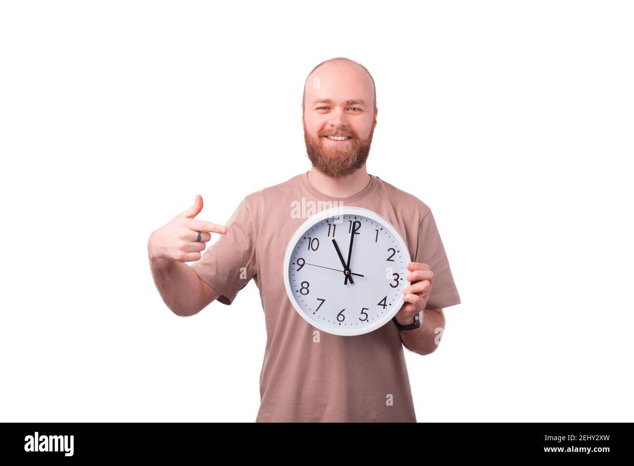 Cheerful smiling young bearded man pointing at white wall watch. Stock Photo