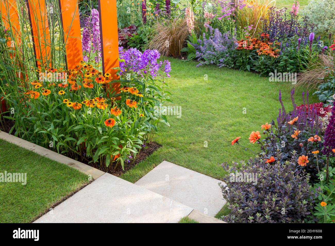 A small English garden - stone steps leading to a lawn grass with flowerbeds filled with flowers plants including Helenium - Dahlias England GB UK Stock Photo