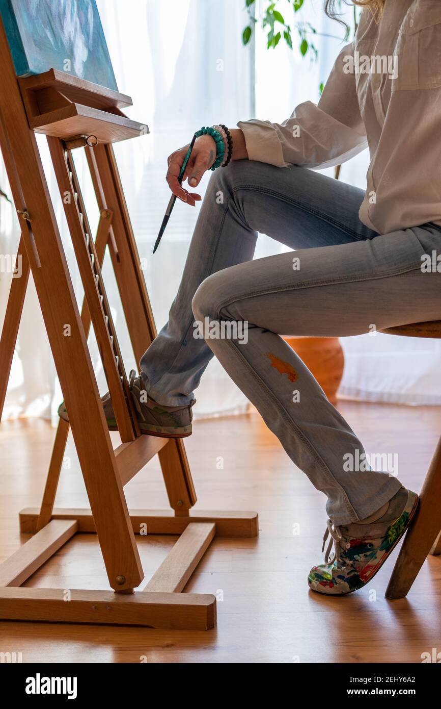 Woman sitting on chair before her paintings on easel. Art and craft concept. Female painter working on her canvas at studio Stock Photo