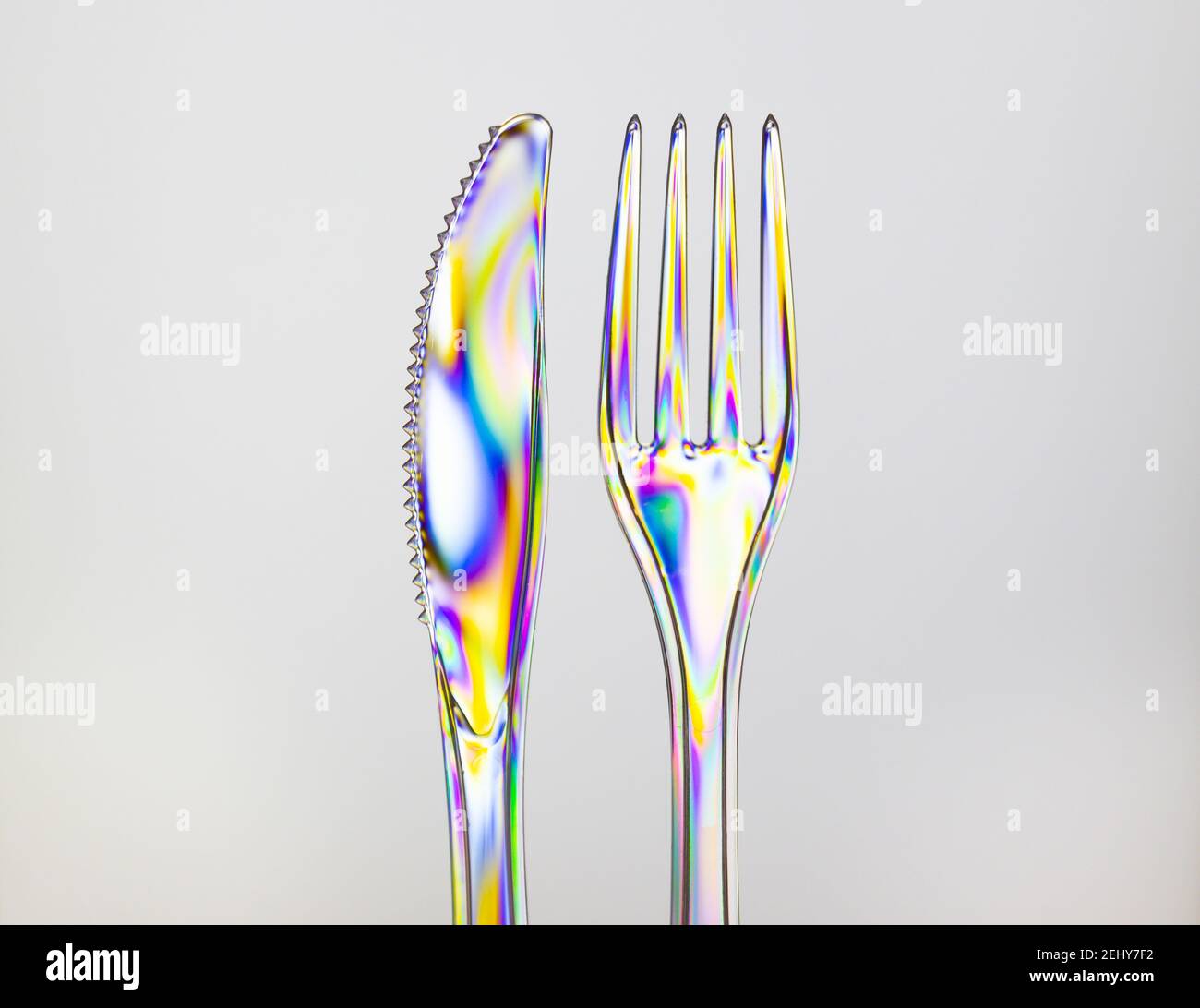 Transparent plastic fork and knife, rainbow colored by photoelasticity isolated on light gray background. Stock Photo