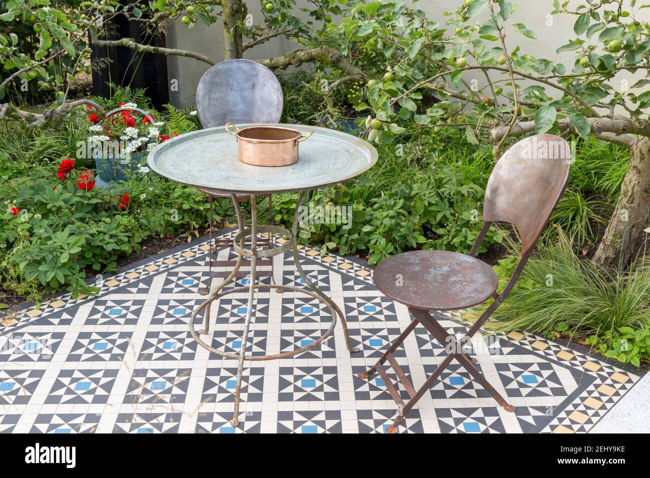 Garden furniture with vintage metal vintage table and chairs on tiled mosaic patio paved paving with apple trees behind a rendered wall England GB UK Stock Photo