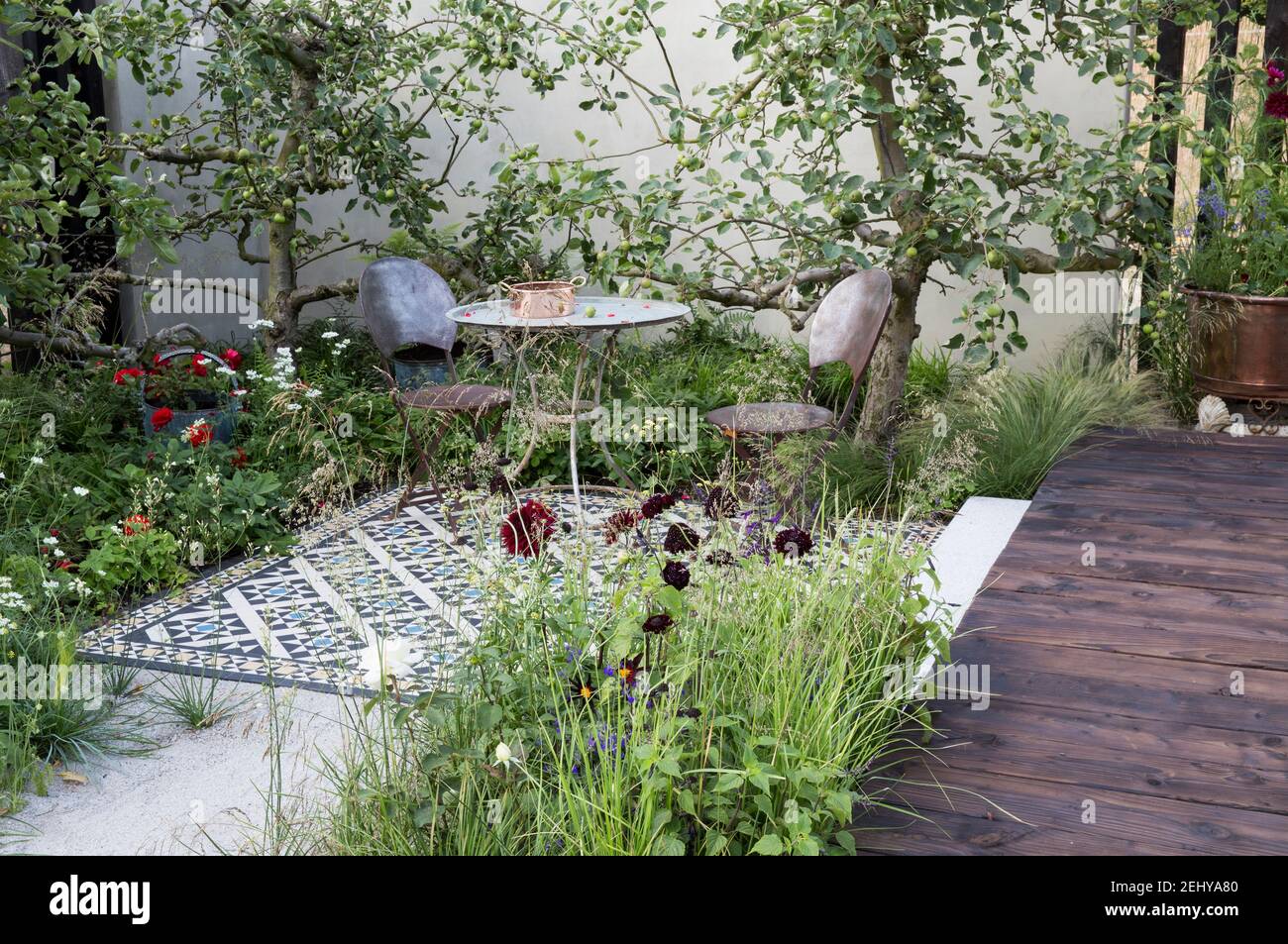 Courtyard garden Table and chairs on tiled mosaic patio with apple fruit trees against a rendered garden wall wooden deck decking in summer England UK Stock Photo