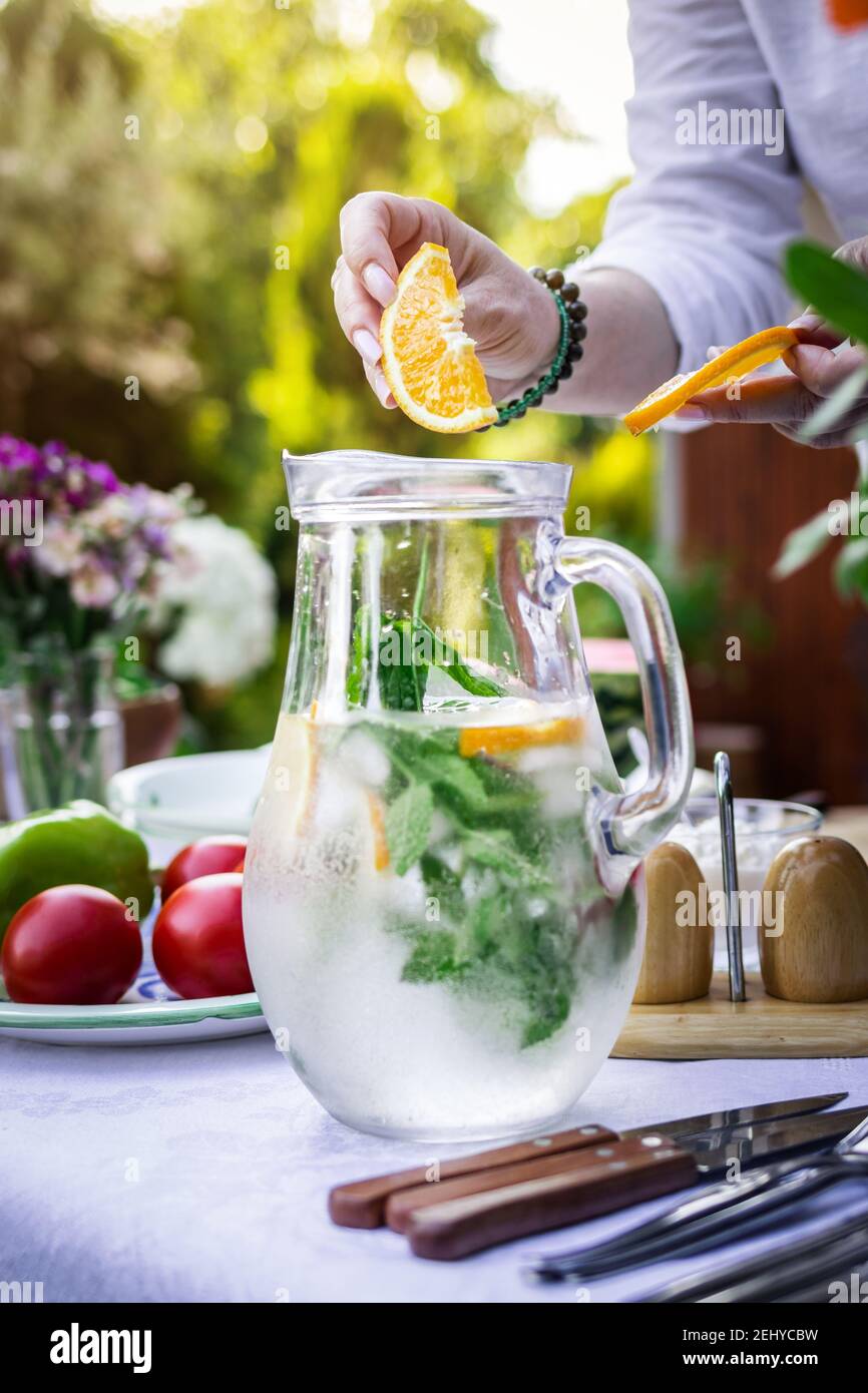 Sparkling water in carafe on table. Woman hand putting slice of orange into cold mineral water with mint and ice Stock Photo