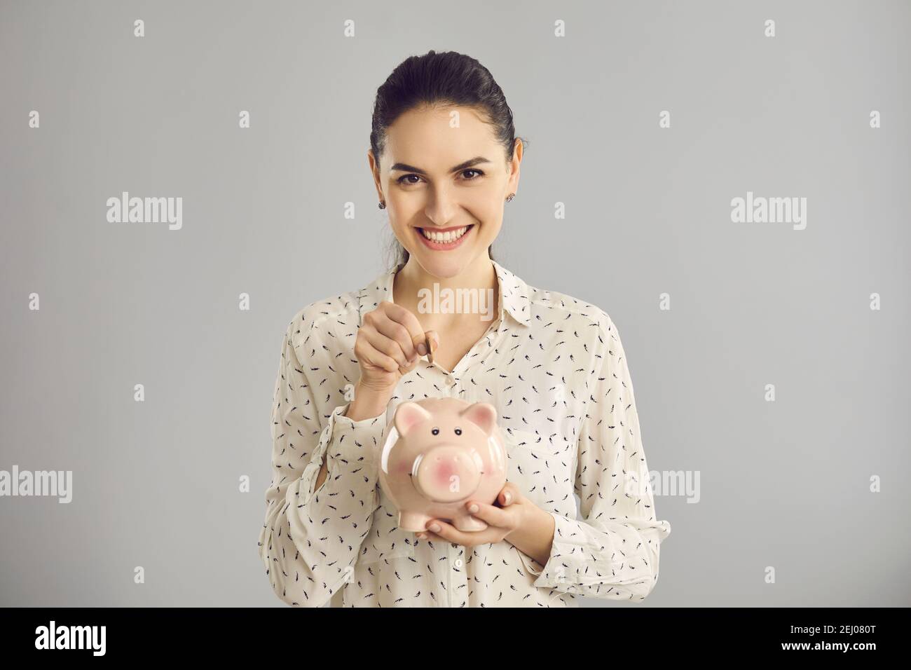 Woman looks at the camera and puts a coin in a piggy bank while standing on a gray background. Stock Photo