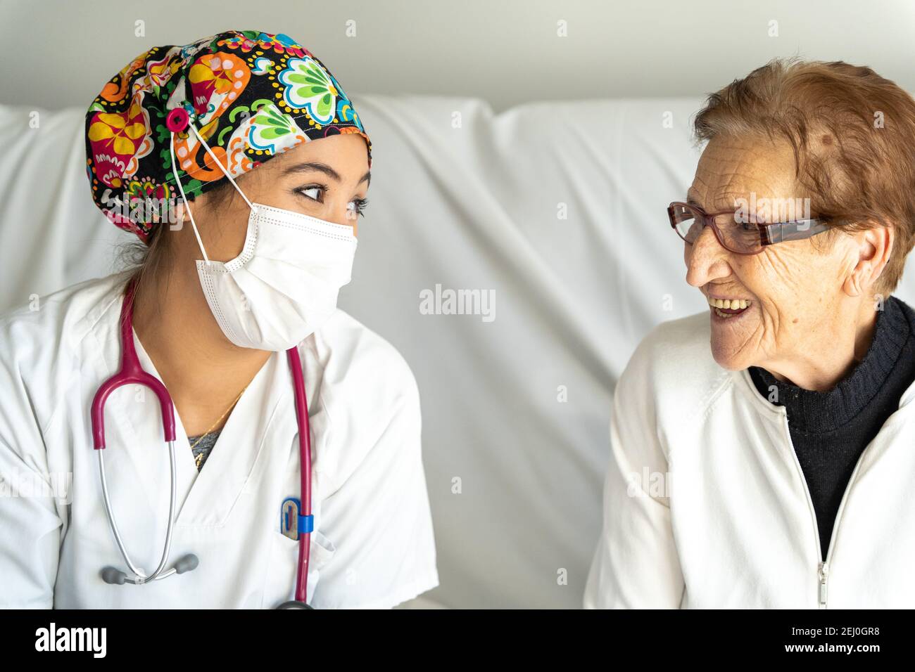 Smiling medic in protective mask holding hand of delighted elderly female while sitting on sofa in apartment and looking at each other Stock Photo