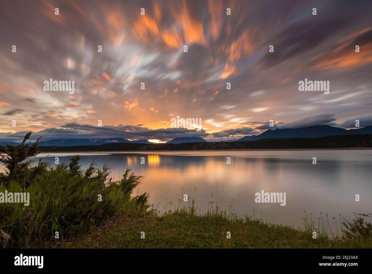 Long exposure calm sunrise from a lake with reflections on water during spring, with orange clouds and mountains on the background, Lake Koocanusa, BC Stock Photo
