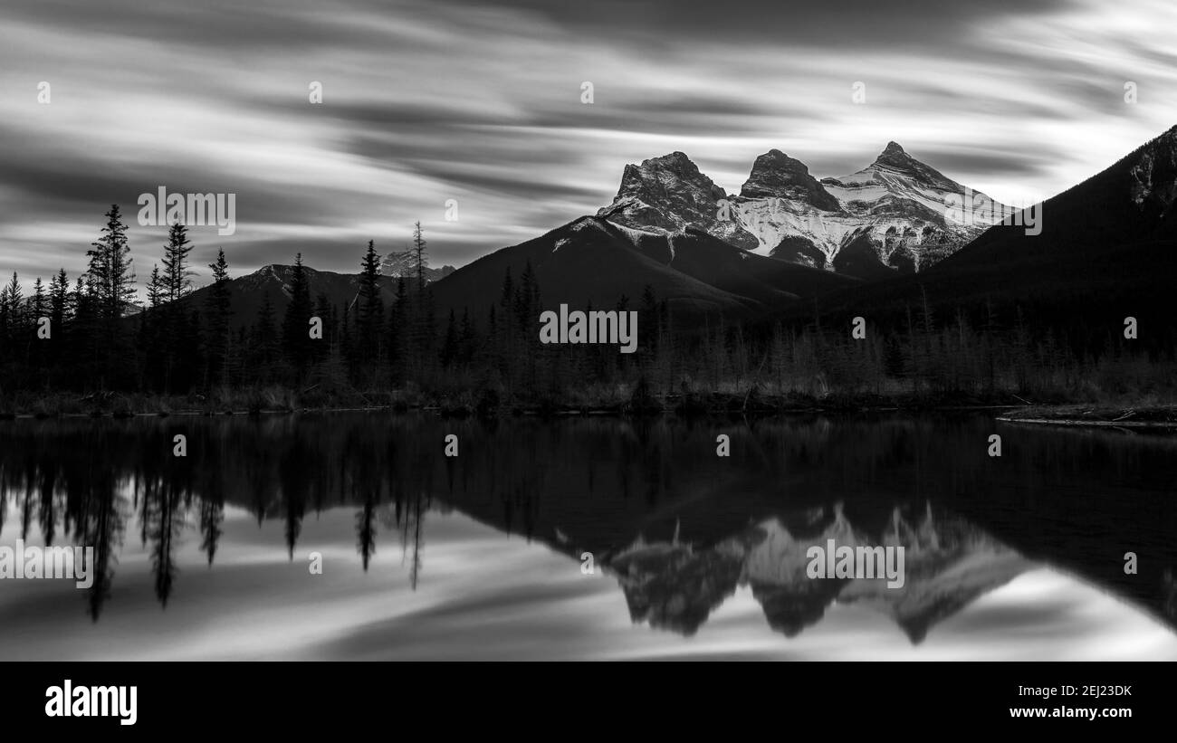 Black and white long exposure of three mountains (Three Sisters) with snow, reflected on a pond under a grey sky with fast clouds during fall, Canmore Stock Photo