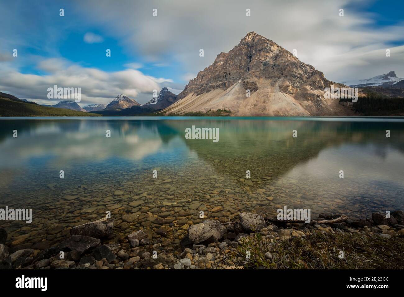 Rocky mountain reflected on clear smooth lake water with green and blue tones and rocks on a summer day with a blue sky and clouds, Alberta, Canada Stock Photo