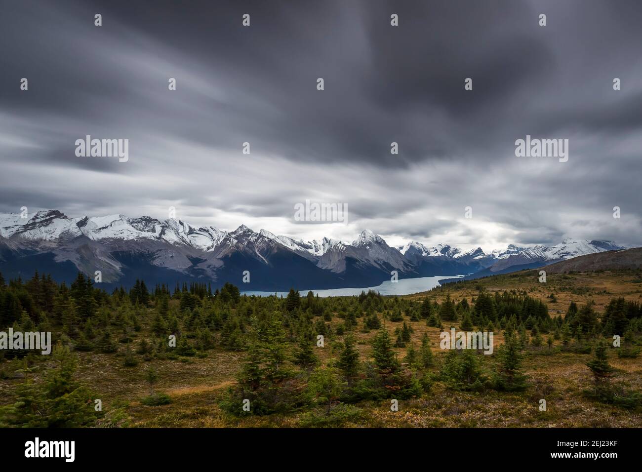 Long exposure of a Canadian Rockies landscape with a lake next to mountains with snow during autumn under a looming overcast sky with fast dark clouds Stock Photo