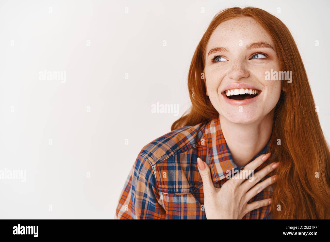 Close-up of beautiful woman with red hair and pale skin laughing happy, holding hand on heart and looking left at logo, standing over white background Stock Photo