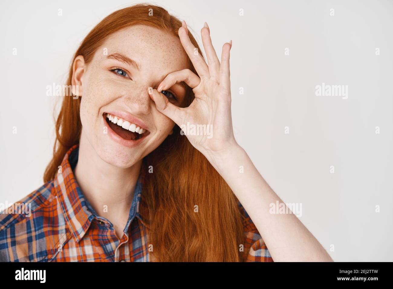 Close-up of happy redhead woman with pale skin and white teeth, showing okay sign on eye and smiling, standing against white background Stock Photo