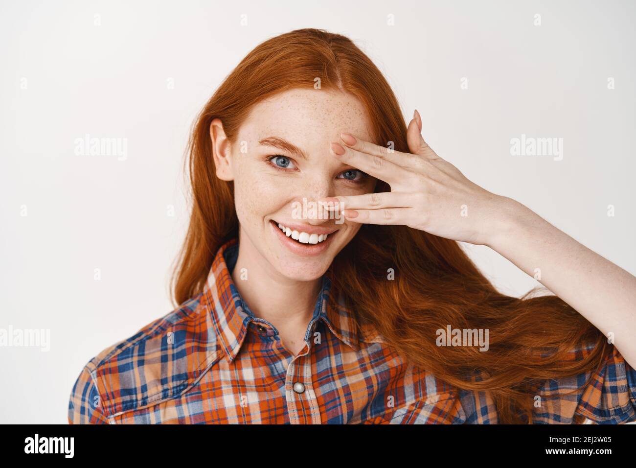 Close-up of beautiful redhead lady with blue eyes and pale skin, smiling at camera, standing over white background Stock Photo