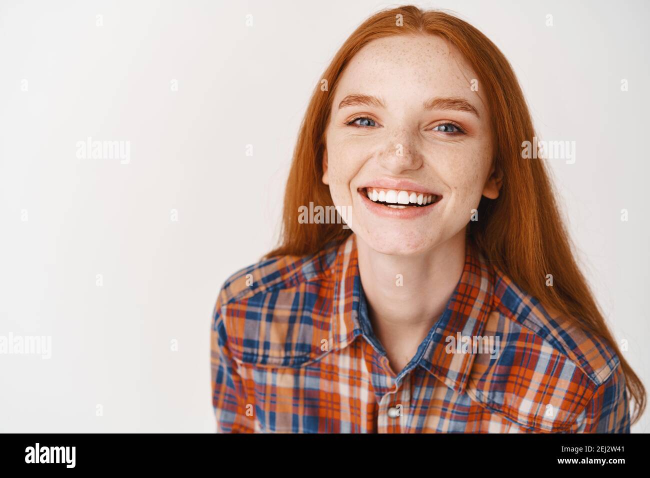 Close-up of beautiful redhead female model with pale skin and white teeth, smiling happy at camera, standing over white background Stock Photo