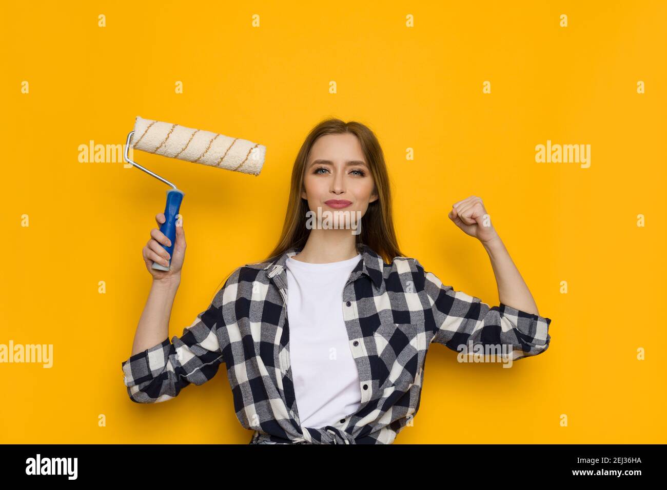 Confident casual young woman in lumberjack shirt is holding paint roller in one hand and other up with fist clenched. Waist up studio shot on yellow b Stock Photo
