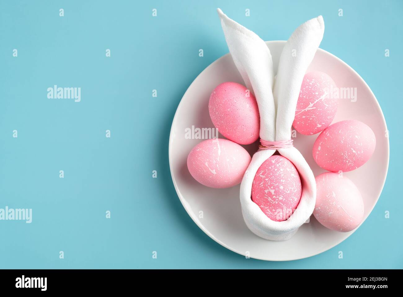 Stylish Easter table setting with easter bunny napkin and pink eggs on plate on turquoise background. Flat lay, top view. Stock Photo