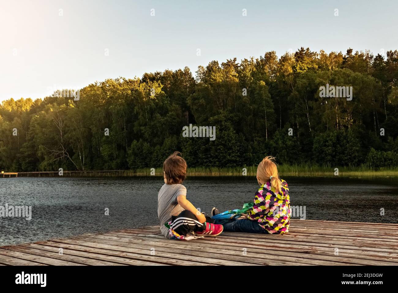 Two little girls are sitting on a wooden pier on the lake at sunset in ...