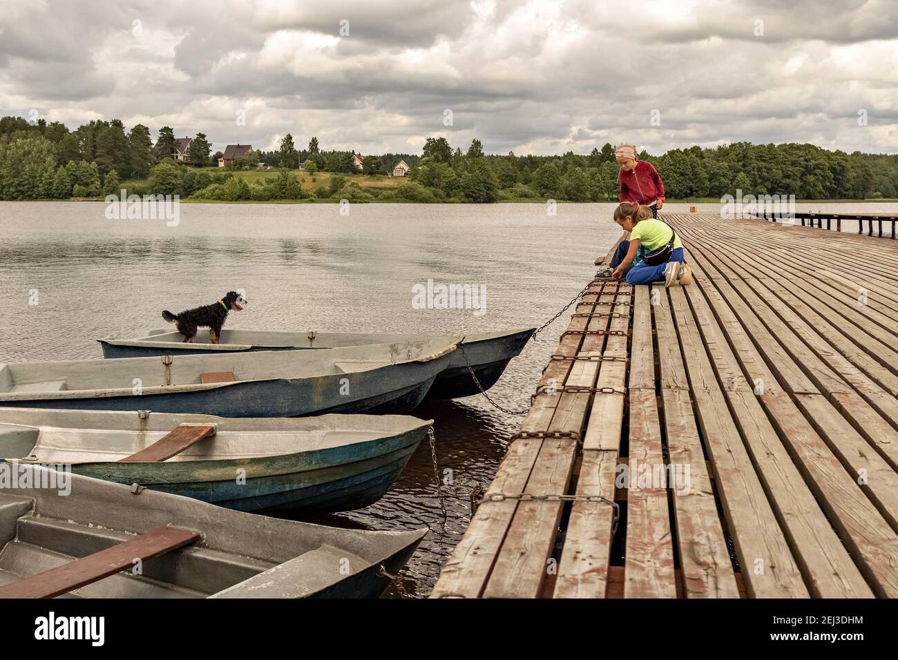Children tie a boat to a wooden pier on a lake at sunset in summer. Dog in the boat. The clouds are thunderous in the sky. Landscape. Stock Photo