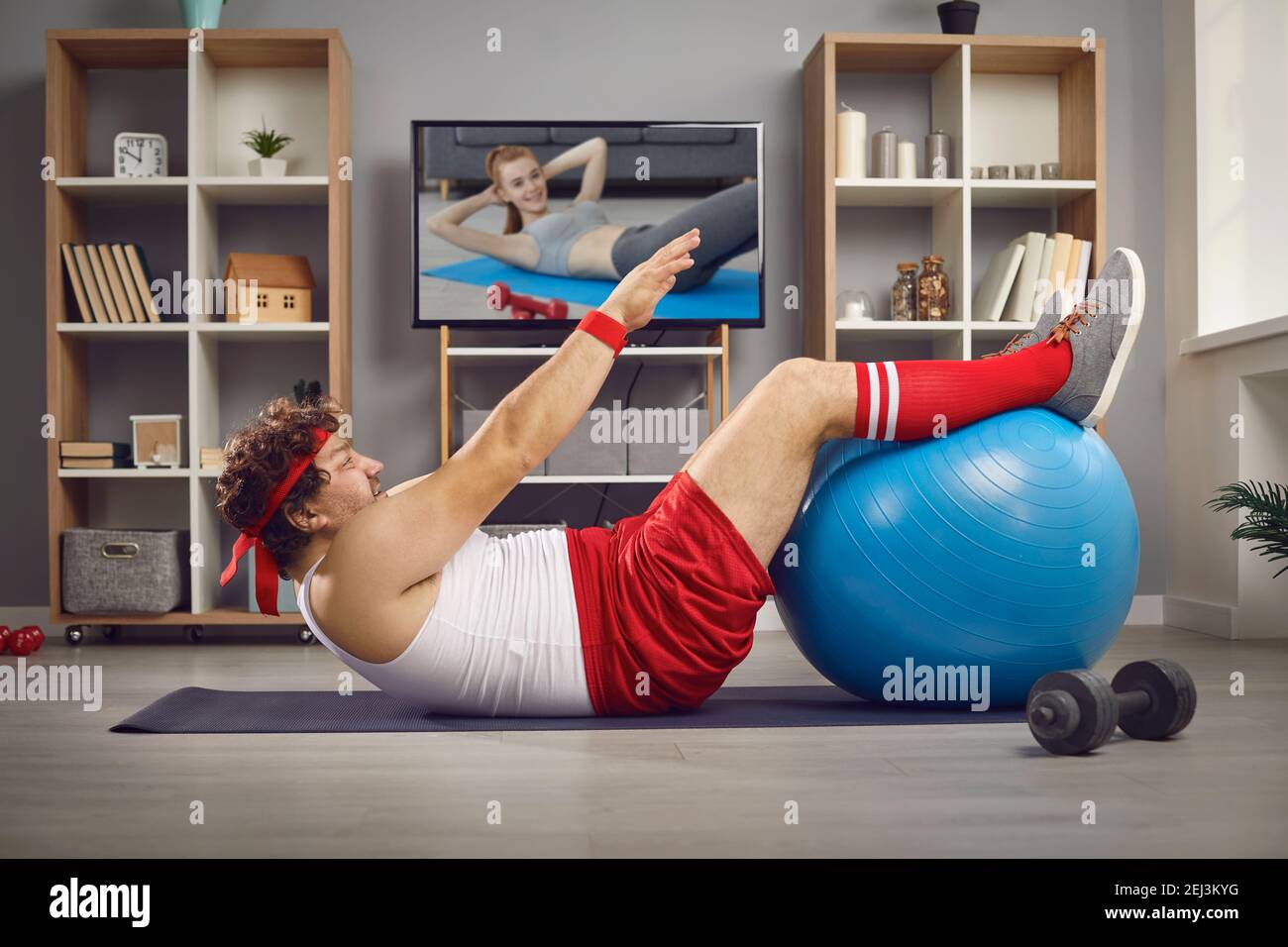 Funny fat man watching TV sports lesson and doing physical exercise with fitness ball Stock Photo