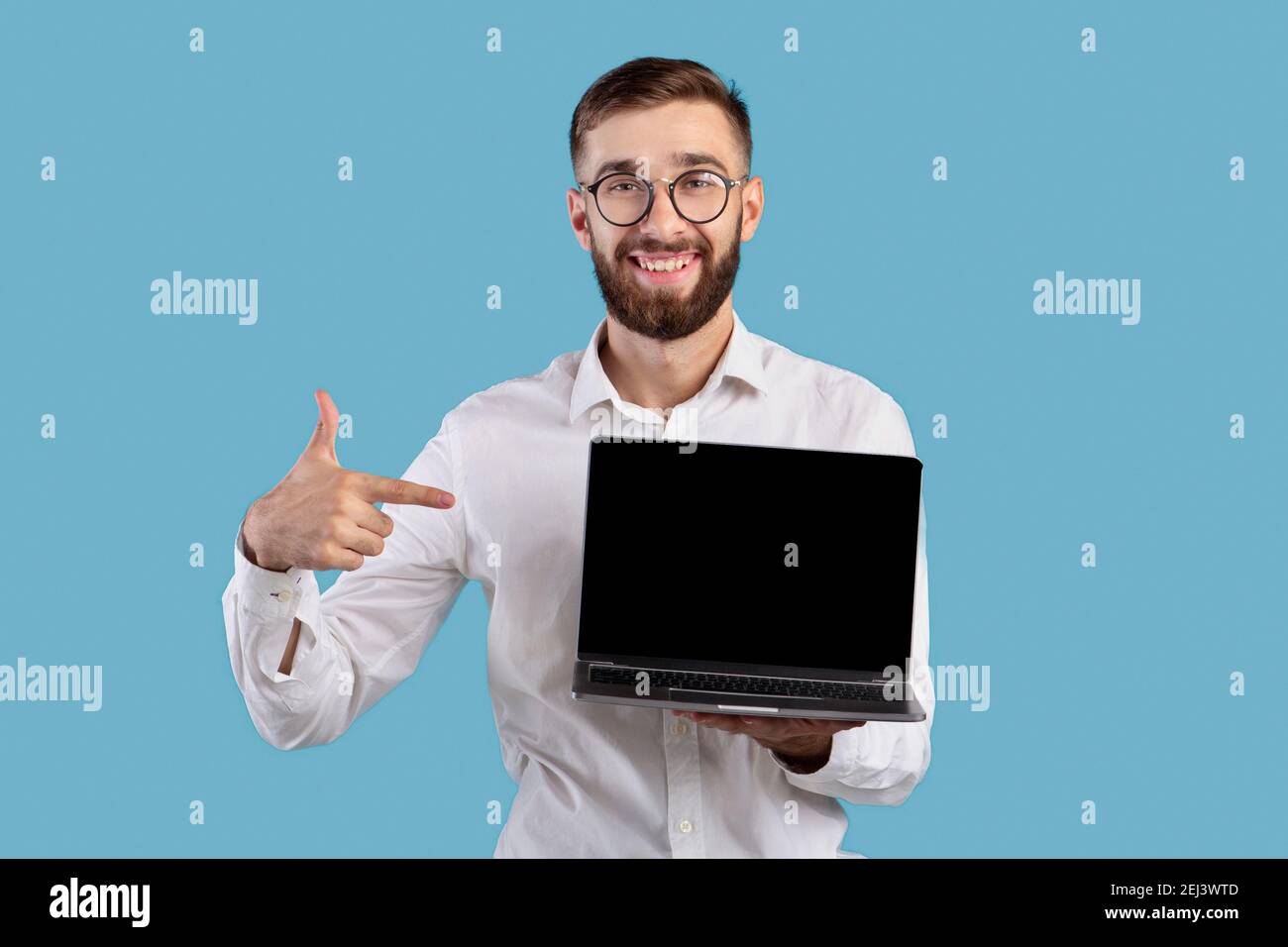 Positive young entrepreneur pointing at laptop computer with blank screen for website design on blue studio background Stock Photo