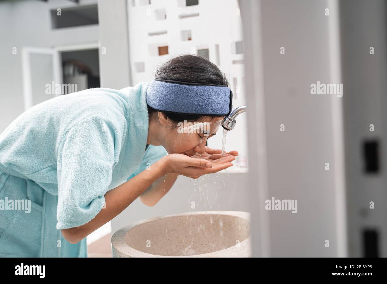 an asian woman wearing a towel wash her face with water in the sink in