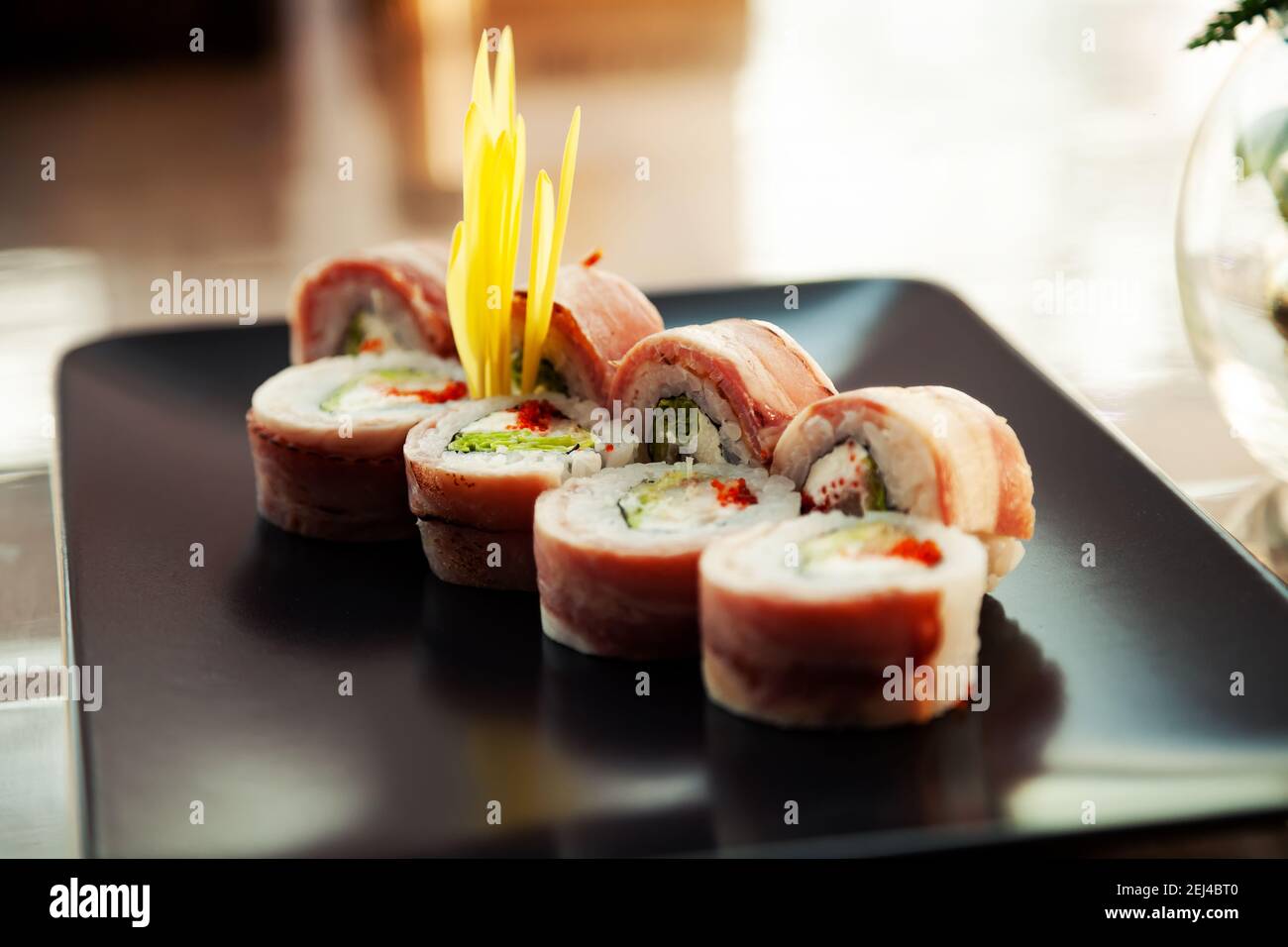 Sushi roll with bacon and fried sea bass on a black plate, ingredients fried sea bass, cream cheese, bacon, Iceberg salad, flying fish roe, Teriyaki sauce, rice, nori. For the restaurant menu Stock Photo