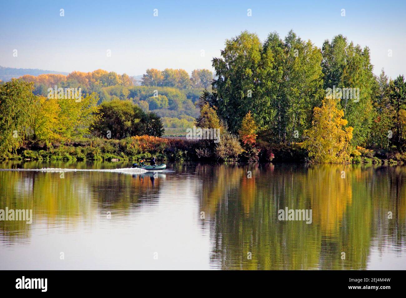 Calmness river surface disturbs driving motor boat. Wonderful landscape of autumn forest on the background. Stock Photo
