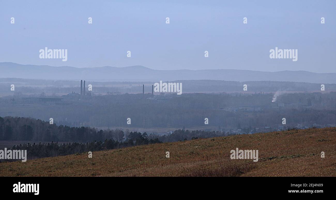View from a hill on a hilly area in a blue haze. You can see the protruding pipes of factories and white smoke. Stock Photo