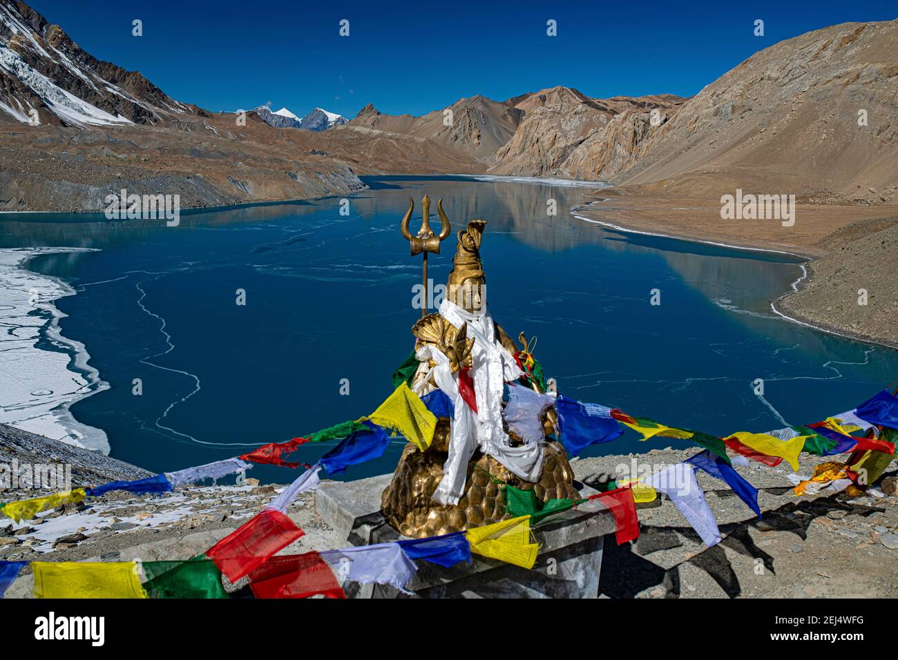Tilicho Lake covered with ice in front of mountain scenery with Buddhist prayer flags and Hindu Shiva statue, often called the highest lake in the Stock Photo