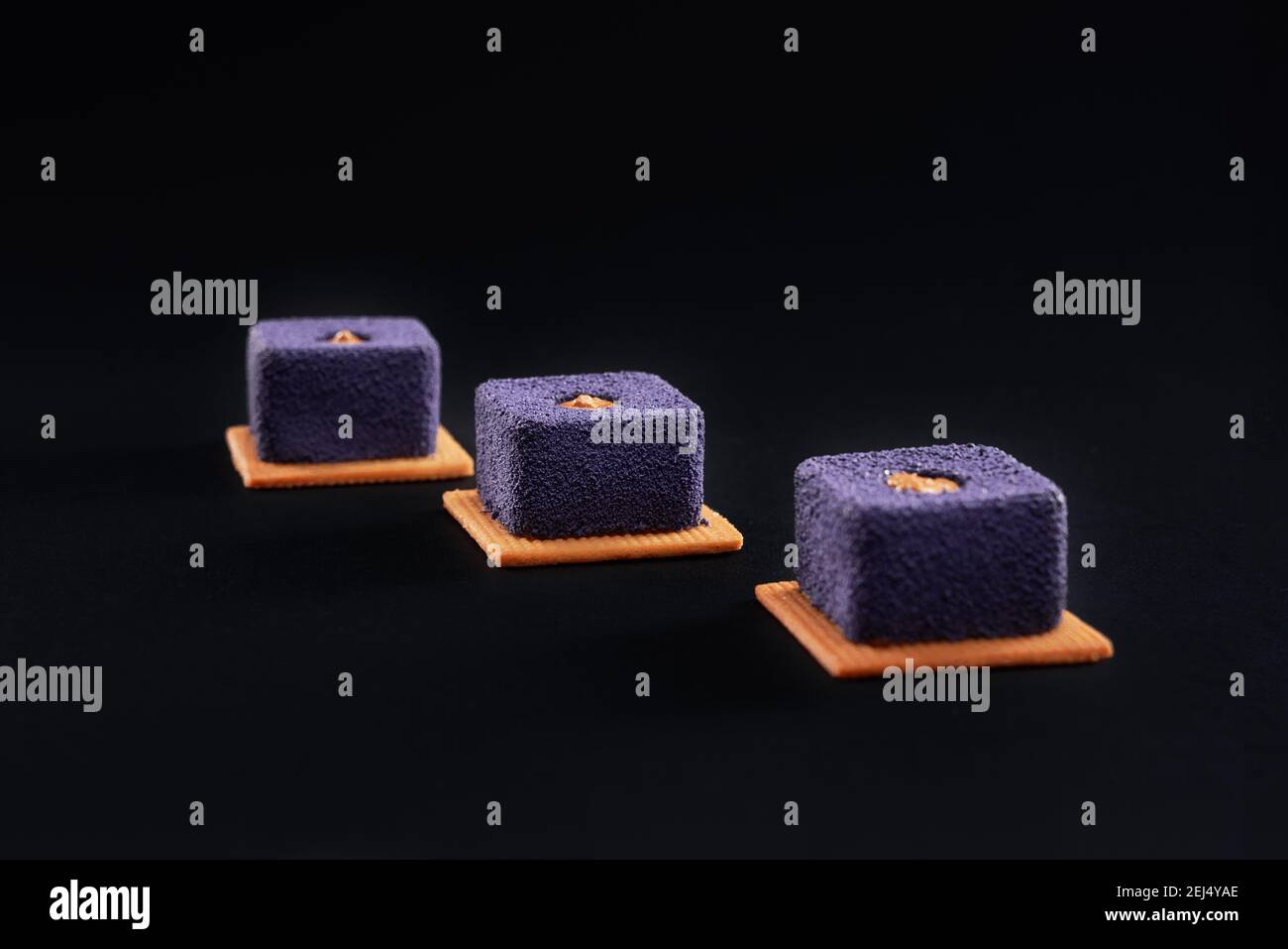 Closeup view of matte purple crunchy dessert filled with brown creme. Three small square matte cakes in row on cookies in restaurant isolated on black Stock Photo