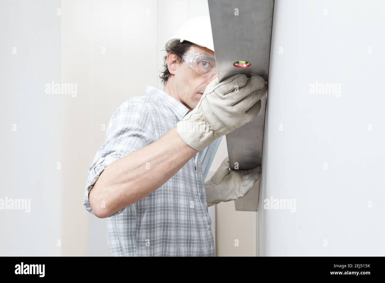 Man drywall worker or plasterer checking level of white plasterboard wall with bubble level at construction site. Wearing white hardhat, work gloves a Stock Photo