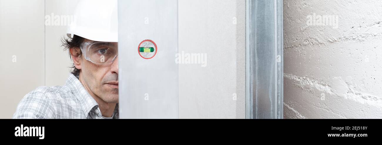 Man drywall worker or plasterer checking level of white plasterboard wall with bubble level at construction site. Wearing white hardhat and safety gla Stock Photo