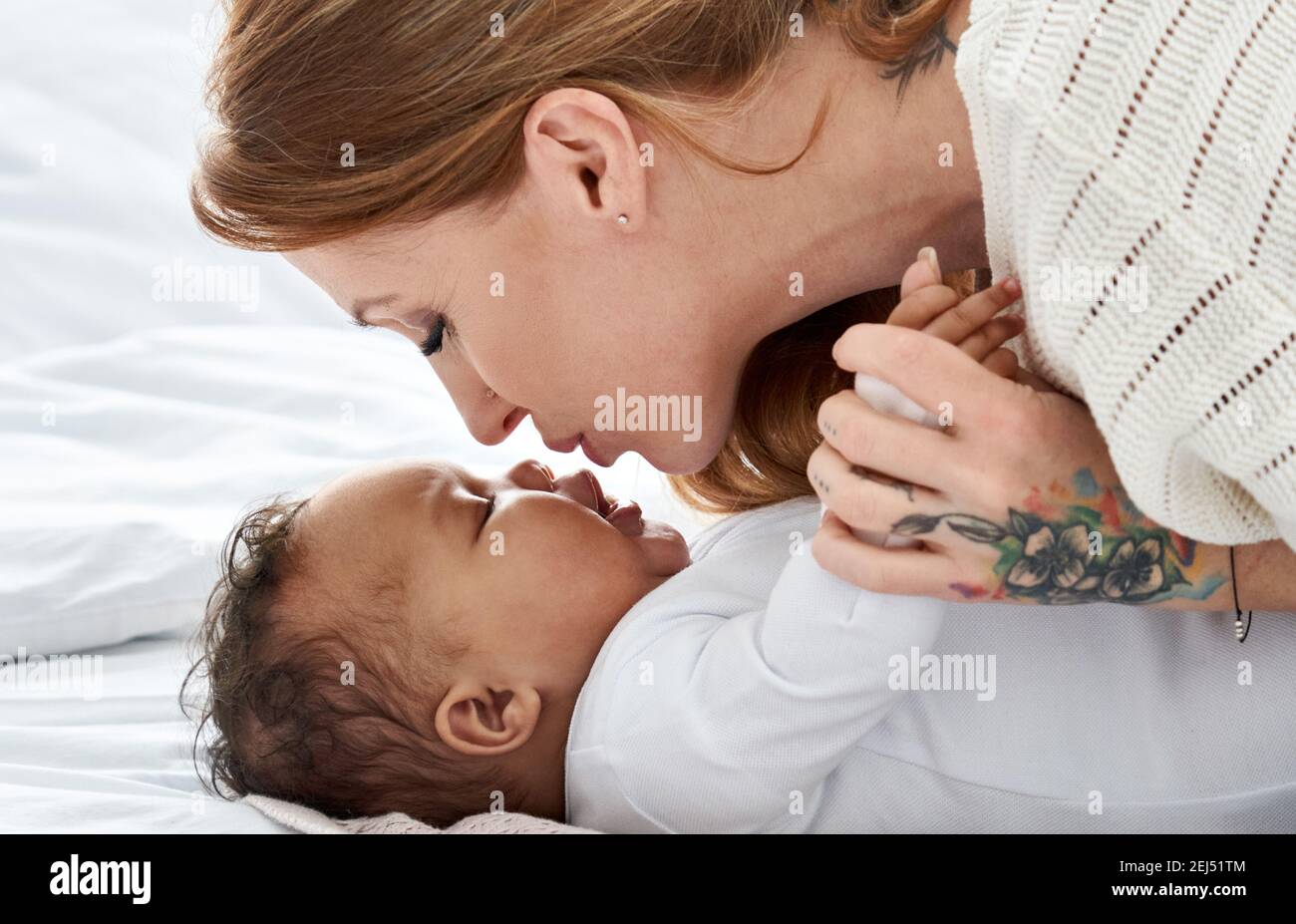 Young caucasian mother kissing cute african american baby daughter lying in bed. Stock Photo