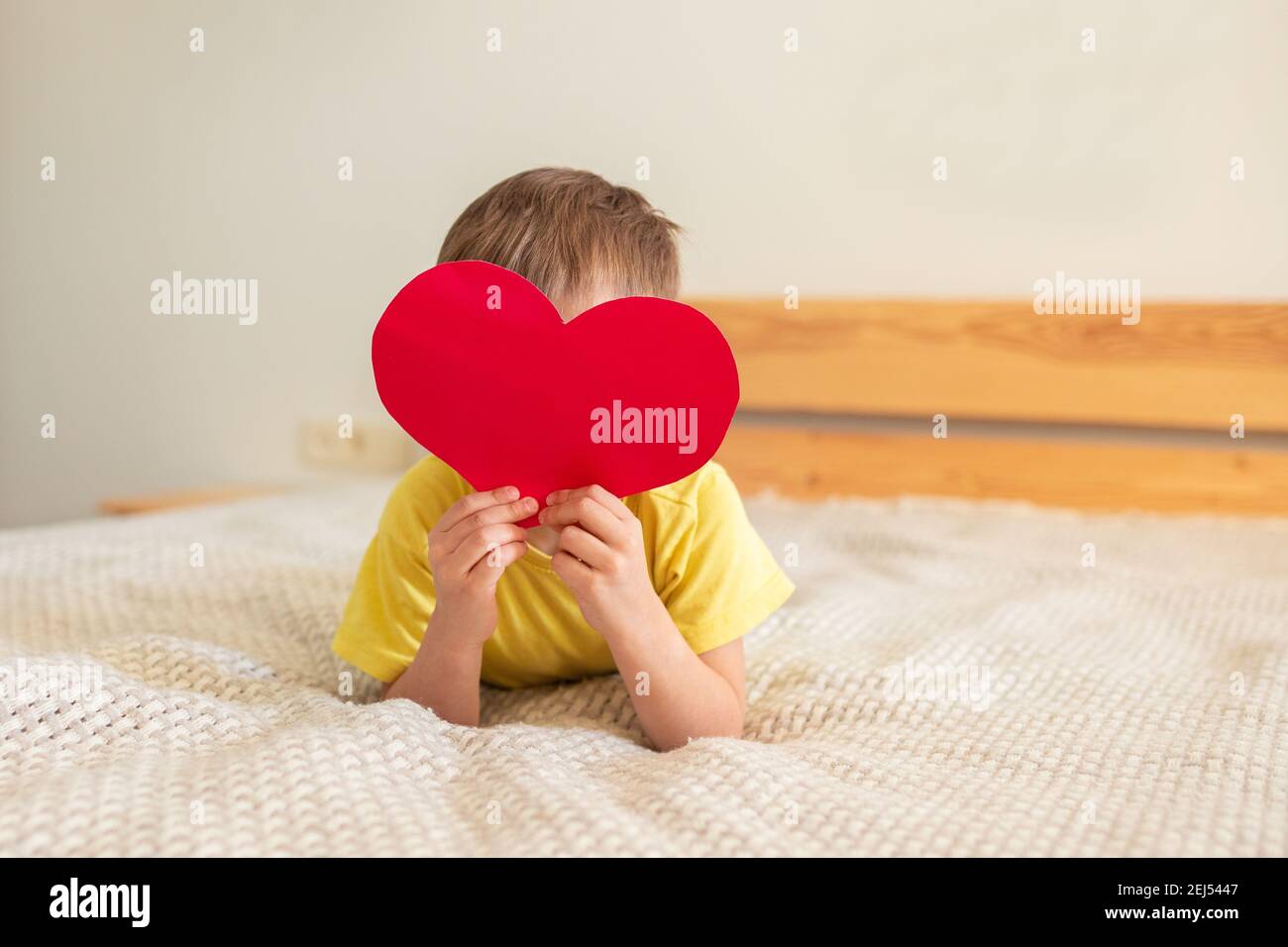 Little boy lying on the bed and holding a red heart made of colored paper, covering his face. Concept for March 8, Mother's Day. Postcard Stock Photo