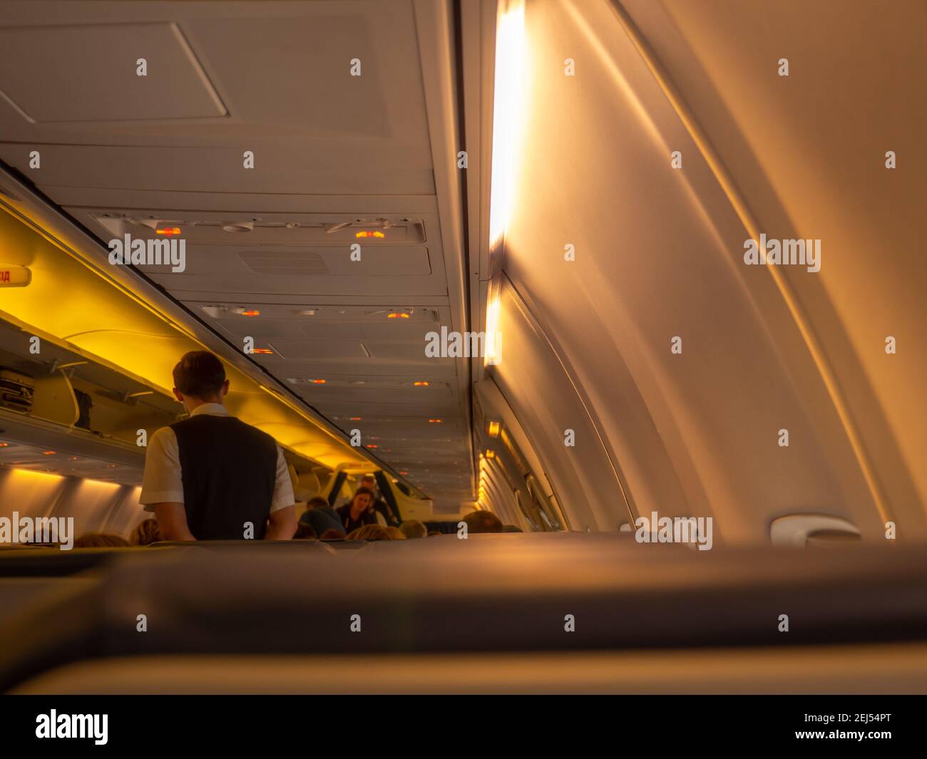 Stewart standing in the aisle of an airplane aircraft cabin. Overhead baggage rack opened and yellow lights turned on. View from the passenger seat. Stock Photo