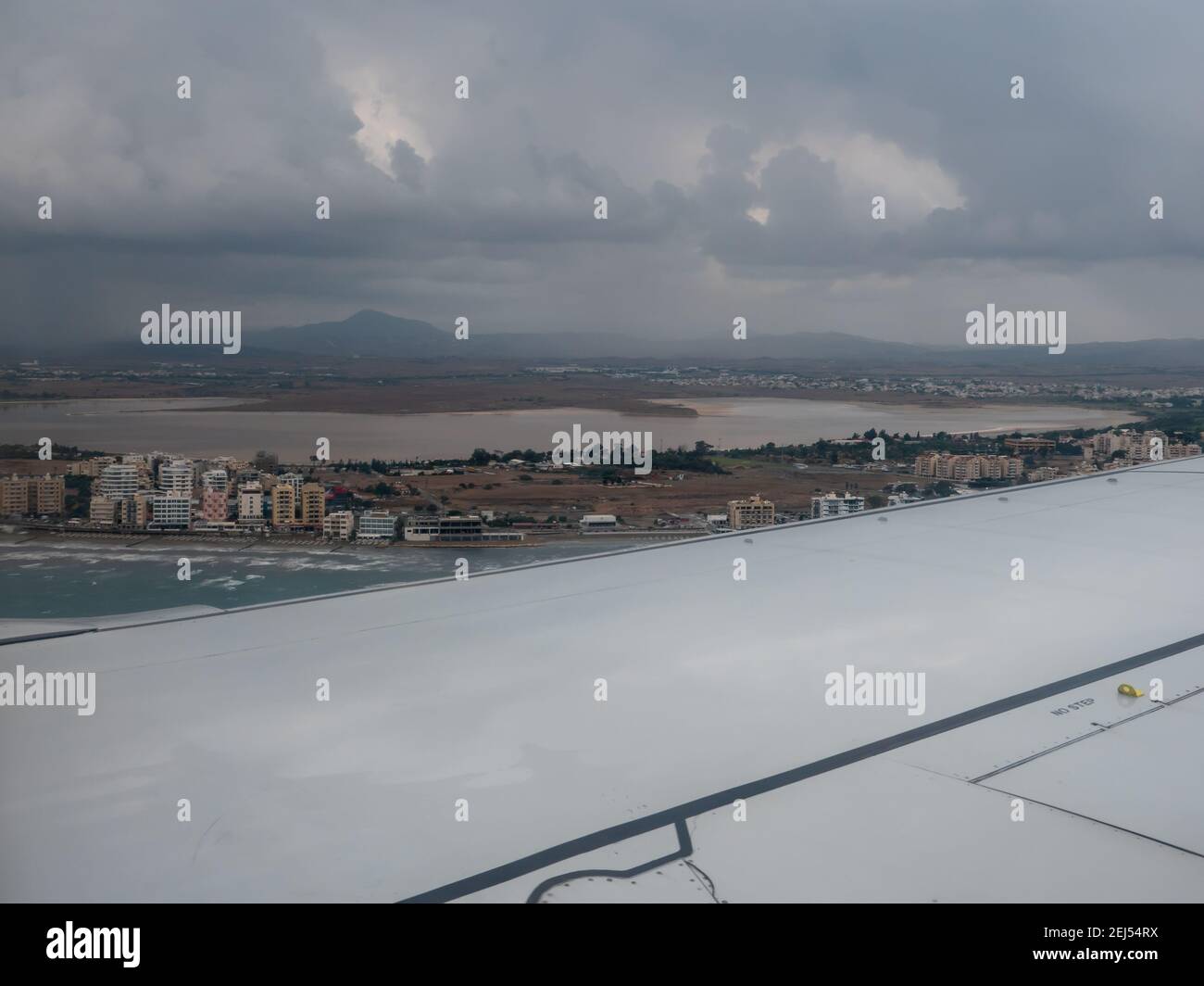 Beautiful view through the airplane window overlooking Larnaca city with buildings and promenade, salt lake, blue sea with waves and mountains. Stock Photo