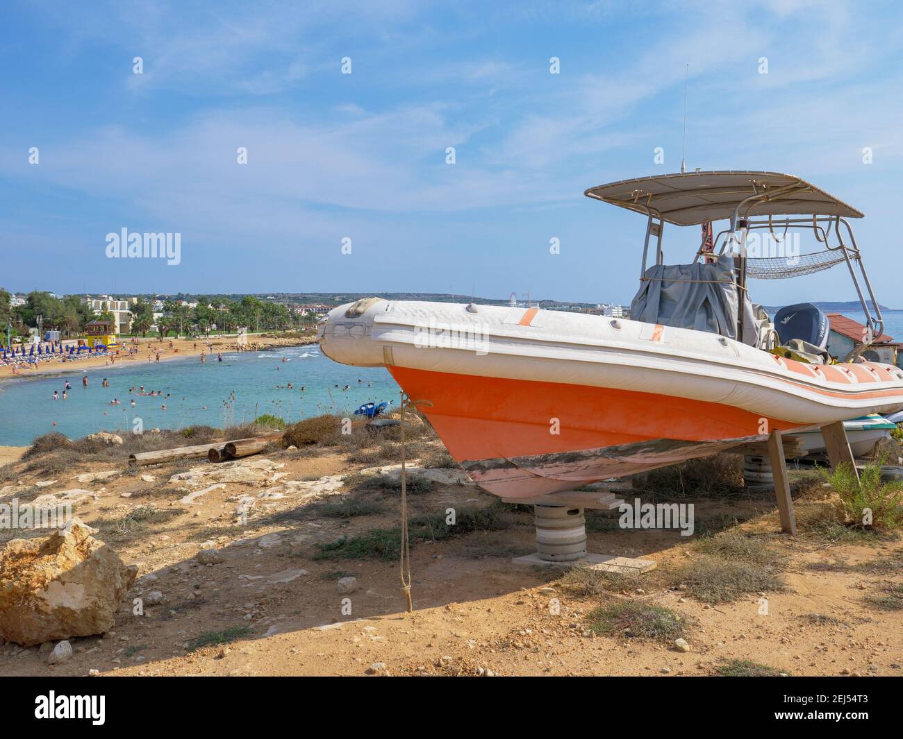 Orange and white powerboat standing on tires and wooden supports at the shore in Ayia Napa. Mediterranean sea in the background. Stock Photo