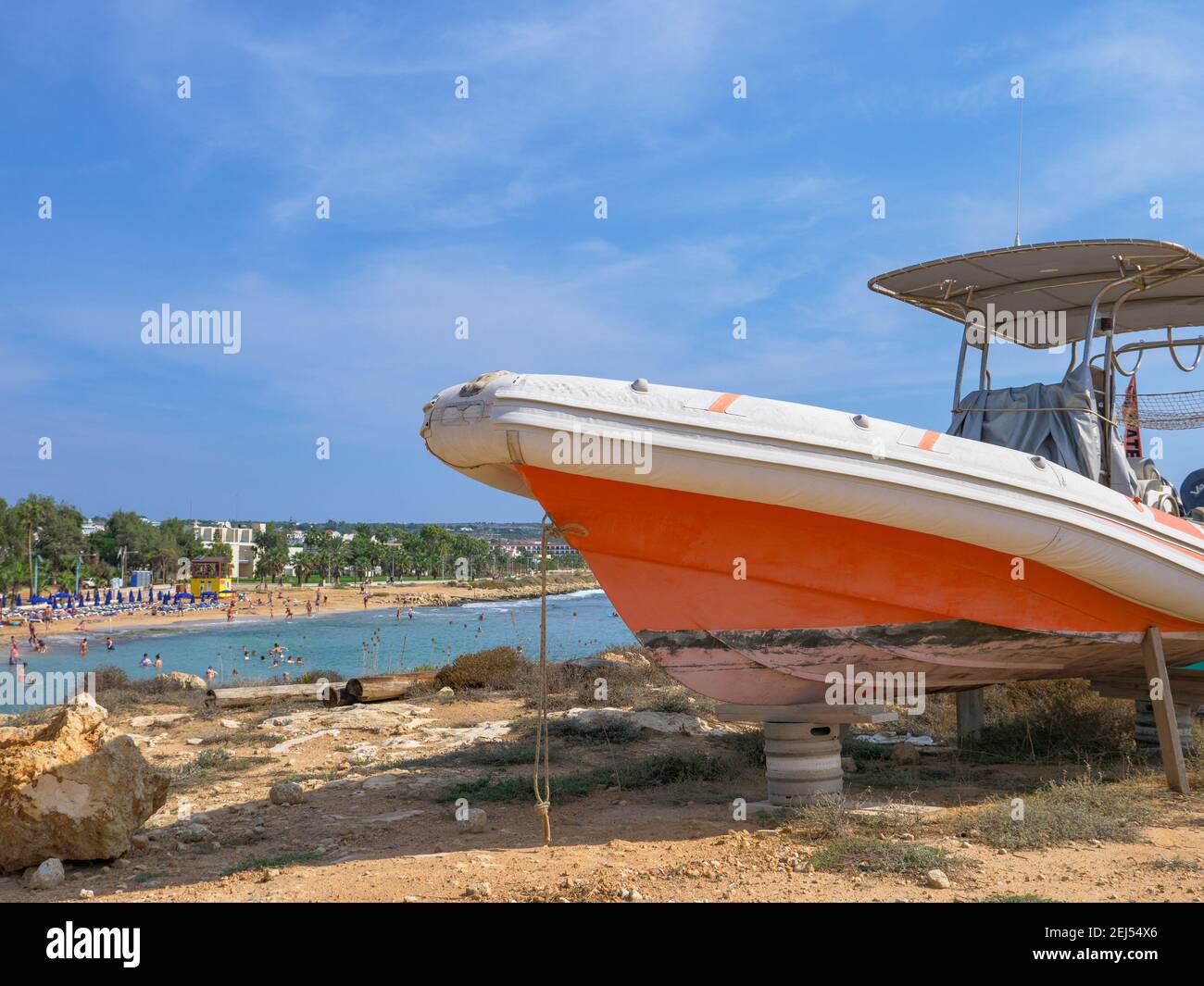 Orange and white powerboat standing on tires and wooden supports at the shore in Ayia Napa. Mediterranean sea in the background. Stock Photo